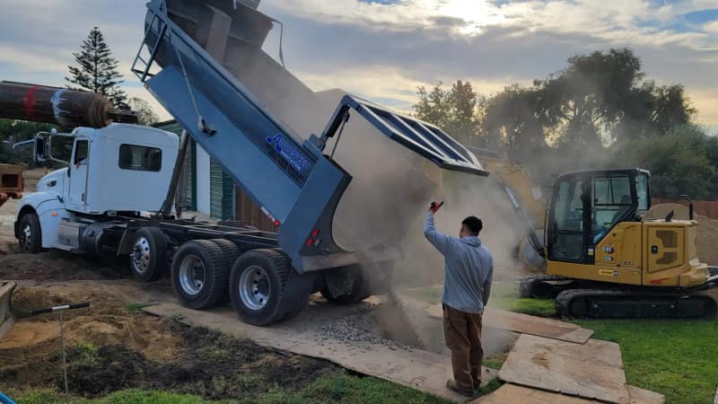 Dump truck unloading fill beside an excavator at a septic installation
