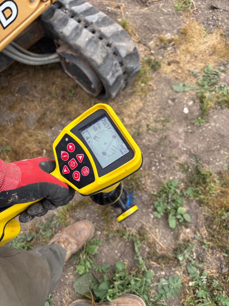 Gloved hand holding a yellow electronic locator over dry ground near a tracked machine