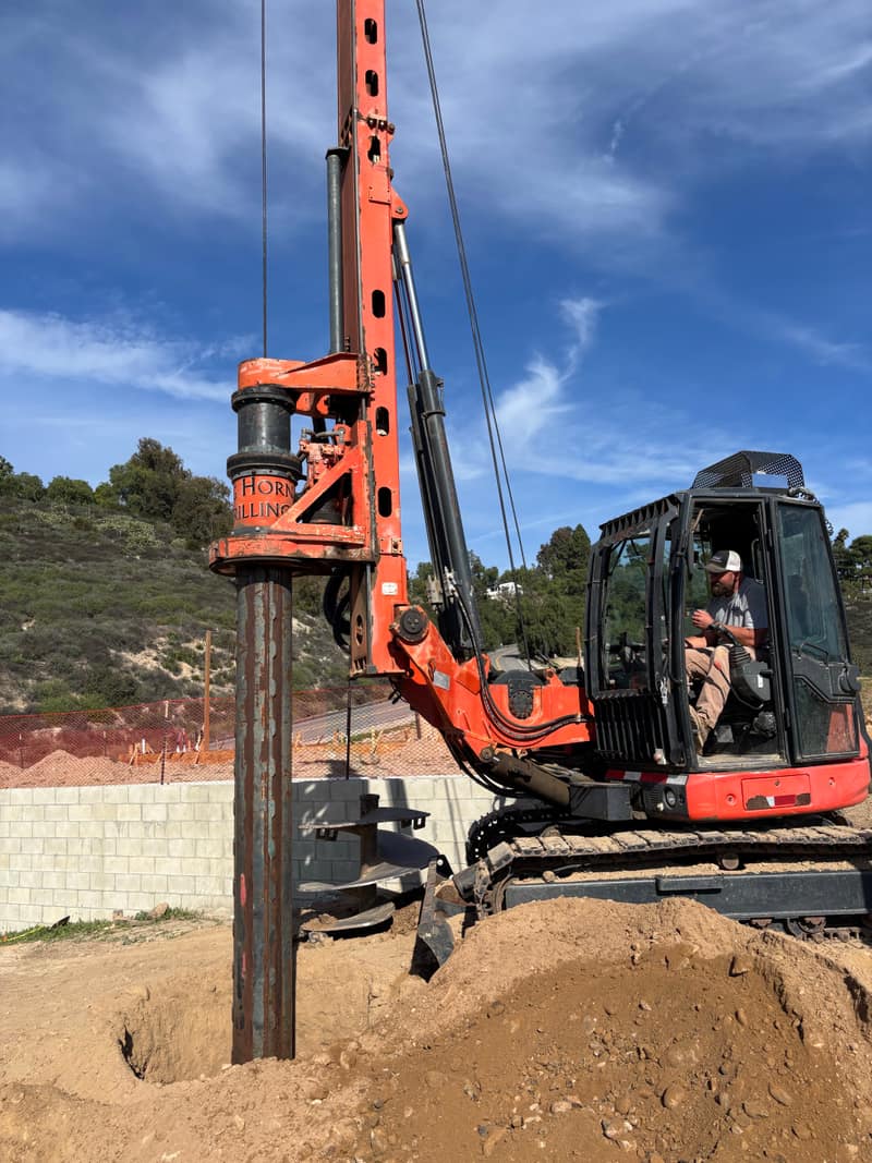 Orange drill rig boring a new seepage pit beside a block retaining wall